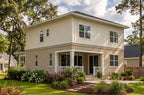 A two-story coastal-style house with beige and cream vertical siding, white trim, a front porch with white columns, and a landscaped lawn with flowers and trees.