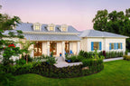 A modern single-story house with a white exterior, a gray metal roof, blue shutters, and a landscaped garden lawn at dusk.